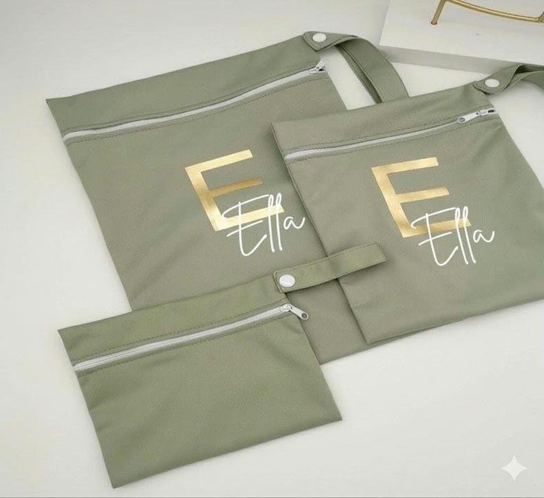 May include: Three sage green zippered pouches of varying sizes. The largest has a handle. Each pouch features the name "Ella" in a gold letter "E" and script font. The zippers are white.