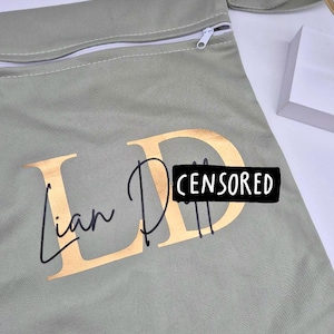 May include: A sage green zippered pouch with gold lettering that reads "Ian D" and a black rectangle that says "CENSORED". The pouch has a white zipper and a small fabric loop with a button closure.