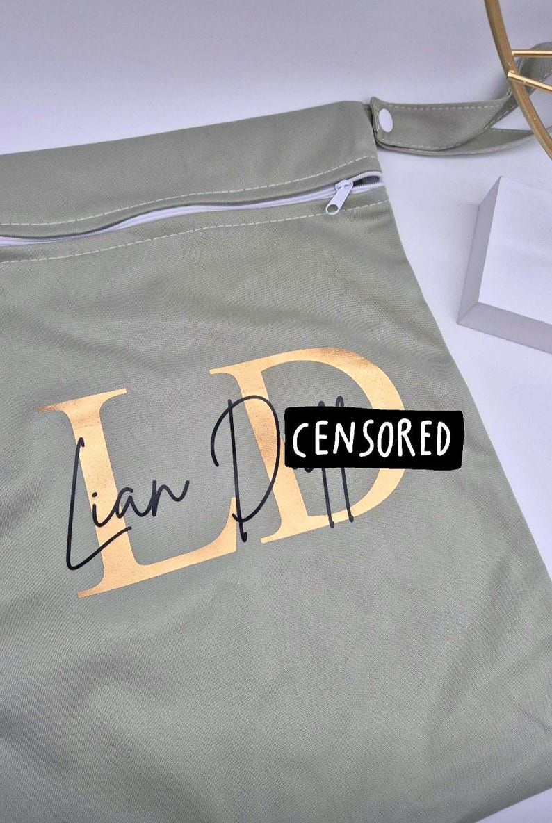 May include: A sage green zippered pouch with gold lettering that reads "Ian D" and a black rectangle that says "CENSORED". The pouch has a white zipper and a small fabric loop with a button closure.