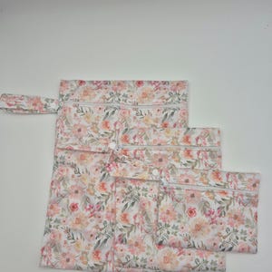 May include: Set of three floral print zippered pouches in varying sizes. The pouches feature a watercolor-style floral pattern in shades of pink, peach, and green on a white background. Each pouch has a white zipper and a matching fabric loop.