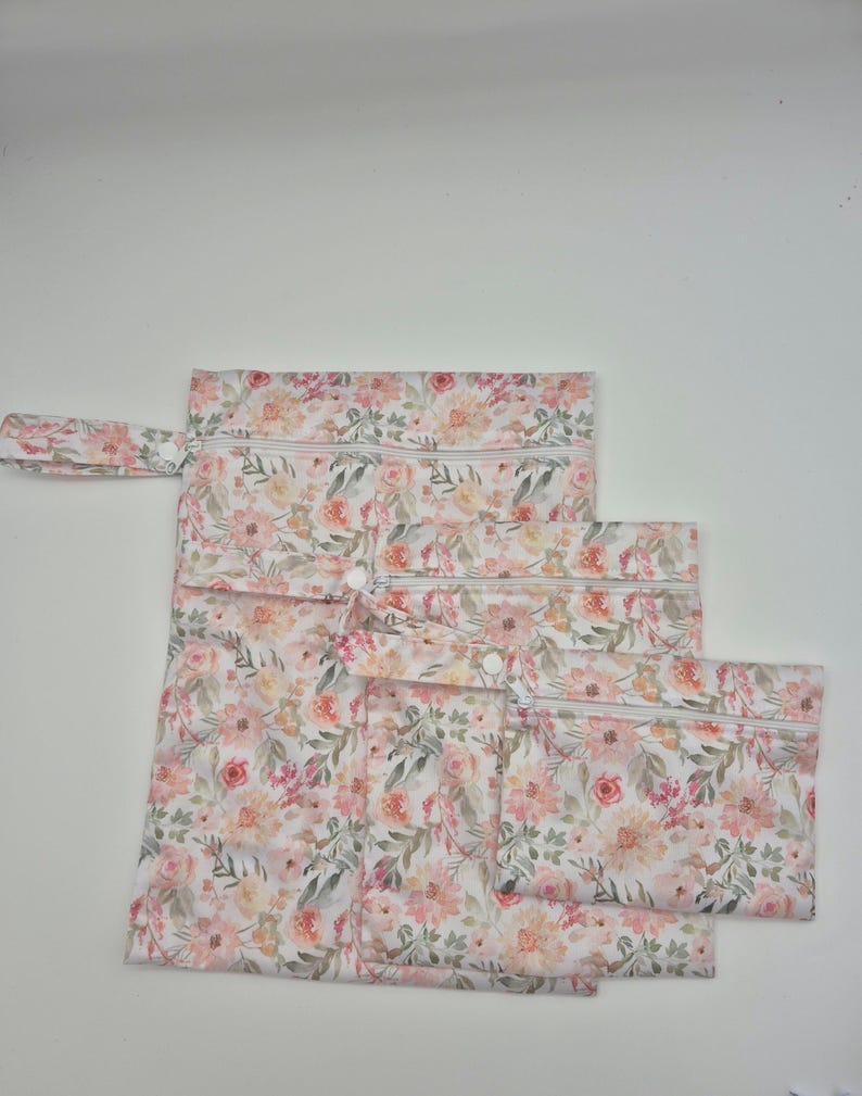 May include: Set of three floral print zippered pouches in varying sizes. The pouches feature a watercolor-style floral pattern in shades of pink, peach, and green on a white background. Each pouch has a white zipper and a matching fabric loop.