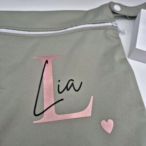 May include: A sage green bag with a white zipper and a snap closure. The bag features the name "Lia" in a pink font with a large pink "L" and a small pink heart.
