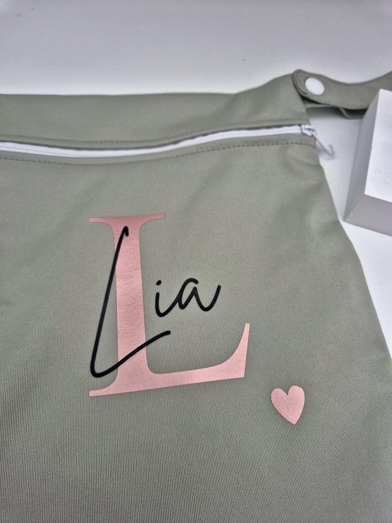 May include: A sage green bag with a white zipper and a snap closure. The bag features the name "Lia" in a pink font with a large pink "L" and a small pink heart.