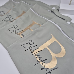 May include: Several sage green zippered pouches with gold foil letters and script text. The pouches have white zippers and a snap closure. The text on the pouches reads "Blubberfische".