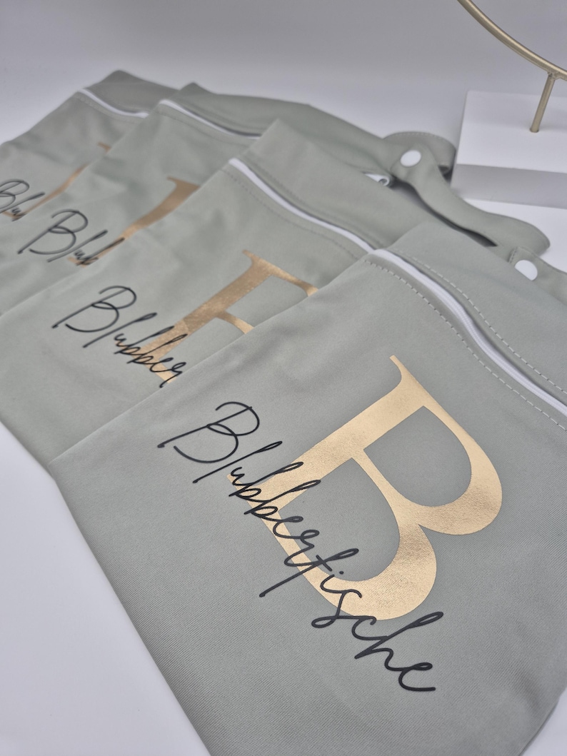 May include: Several sage green zippered pouches with gold foil letters and script text. The pouches have white zippers and a snap closure. The text on the pouches reads "Blubberfische".