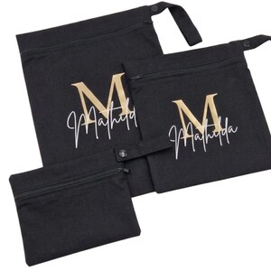May include: Three black fabric bags with zippers and a snap closure. The bags have a gold-colored letter "M" and the name "Matilda" in white script. The bags are in different sizes.