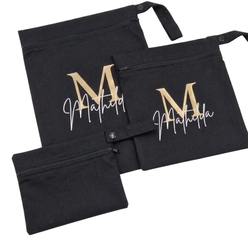 May include: Three black fabric bags with zippers and a snap closure. The bags have a gold-colored letter "M" and the name "Matilda" in white script. The bags are in different sizes.