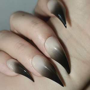 May include: Close-up of long, pointed false nails with a gradient design. The nails transition from a pale beige at the base to a deep black at the tips. The nails are smooth and glossy, creating a dramatic effect.