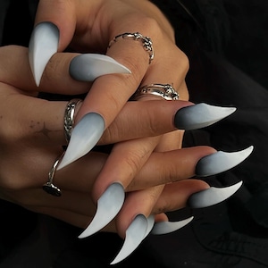 Könnte beinhalten: Nahaufnahme von Händen mit langen, spitzen, ombre-Nägeln. Die Nägel sind in einem Farbverlauf von Dunkelgrau zu Weiß lackiert. Die Nägel sind lang und spitz, mit einer spitzen Spitze.