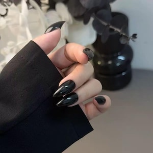 Puede incluir: Uñas de estilete negras brillantes. Las uñas son largas, puntiagudas y tienen un acabado brillante. La mano lleva una manga negra. El fondo incluye elementos decorativos en blanco y negro.