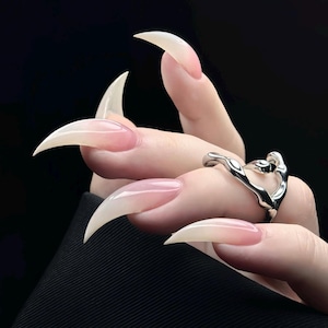 May include: Close-up of long, pointed, clear acrylic nails with a pink ombre effect. A silver ring with an abstract design is on a finger. The background is black.