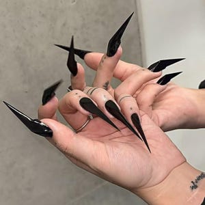 May include: A close-up of hands with long, pointed, black acrylic nails. The nails are glossy and sharp, extending far beyond the fingertips. The hands also have tattoos, including dots and lines on the fingers and a wrist tattoo.