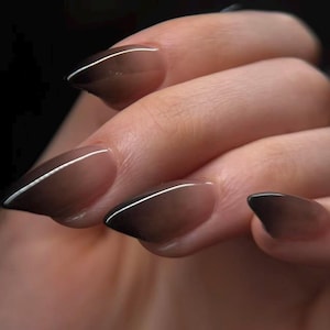 May include: Close-up of a hand with long, almond-shaped nails. The nails have a gradient design, fading from a light brown base to a dark brown tip. A thin white line accents the edge of each nail, creating a modern look.