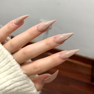 May include: Close-up of long, pointed, beige-colored artificial nails. The nails have a glossy finish and are slightly glittery. The hand is wearing a white knitted sweater.