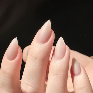 May include: Close-up of a hand with long, pointed, light pink nails. The nails are a natural colour and have a sharp, stiletto shape. The background is a dark, solid colour, highlighting the nails.