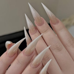 May include: Long, stiletto-shaped artificial nails with a gradient design. The nails transition from a clear base to a light beige color at the tips. The nails are long and pointed.
