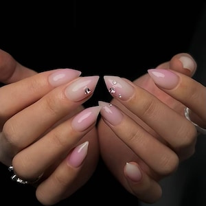 May include: A set of long, stiletto-shaped nails with a gradient pink and white design. Each nail is adorned with small, sparkling rhinestones. The nails are set against a dark background.