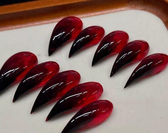 May include: A set of ten long, pointed artificial nails. Each nail features a gradient design, transitioning from a deep red at the base to a black tip. The nails are arranged on a white surface, with a wooden tray in the background.