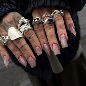 Op de afbeelding: Close-up van handen versierd met meerdere zilveren ringen van verschillende ontwerpen, waaronder een schedel, een getextureerde band en een grote ovale ring. De nagels zijn lang, met een doorschijnende roze tint.