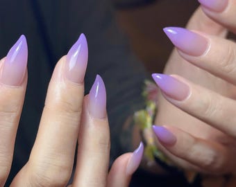 May include: Pointed almond-shaped nails featuring a smooth gradient transition from a clear, natural base at the cuticle to a vibrant, solid lavender purple at the tips. The finish is glossy and reflective, highlighting the sharp, tapered shape.