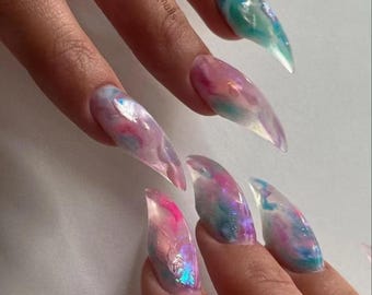 May include: Close-up of long, pointed, clear acrylic nails with a pink, blue, and white marbled design. The nails are decorated with iridescent glitter.
