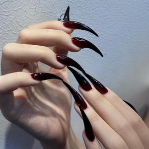 May include: Long, pointed artificial nails with a dark red to black gradient. The nails are highly polished and extend well beyond the fingertips, creating a dramatic and edgy look. The nails are a deep burgundy color at the base, fading to black at the tips.