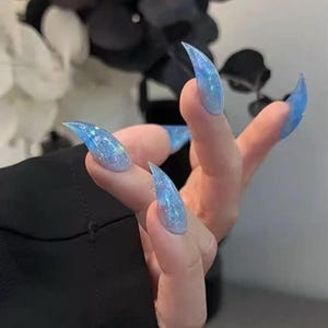 Puede incluir: Una mano con uñas largas y puntiagudas con purpurina azul. Las uñas tienen un aspecto translúcido con purpurina incrustada, creando un efecto brillante. Las uñas son de color azul claro, con purpurina plateada y azul.