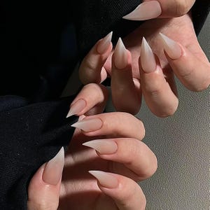 Könnte beinhalten: Nahaufnahme von Händen mit langen, spitzen, klaren, weißen, Ombre-Nägeln. Die Nägel sind an der Basis hell milchig weiß und gehen allmählich zu einem klaren, durchscheinenden Weiß an den Spitzen über.