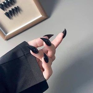 Pode incluir: Unhas postiças pretas e brilhantes em forma de estilete. As unhas são longas e pontiagudas, com um acabamento liso e polido. Um conjunto de unhas semelhantes é visível no fundo. Um acessório arrojado.