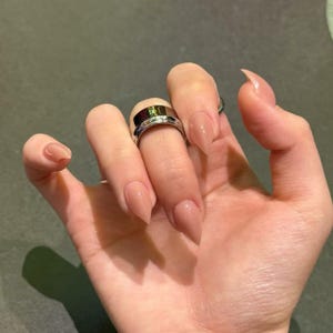 以下が含まれることがあります： ヌードカラーにペイントされた、長くアーモンド型の爪を持つ手。幅広のシルバーリングと細いリングが薬指を飾っています。爪は磨かれ、光沢のある仕上がりで、自然でニュートラルな色合いです。