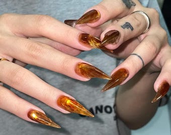 May include: Long, pointed, amber-colored nails with a translucent, marbled effect.