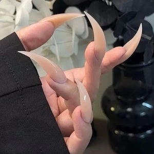 以下が含まれることがあります： 滑らかで光沢のある仕上げの、長く尖ったヌードカラーの人工爪のセット。爪は劇的なスティレット形状をしており、外側に湾曲し、自然な爪床を大幅に超えて伸びる単色で不透明なトーンが特徴です。