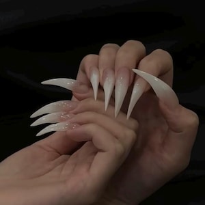 May include: A hand displaying long, pointed, white acrylic nails. The nails have a clear base with a gradient effect, transitioning to white tips. The background is a solid black.