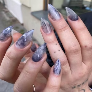 May include: Close-up of long, pointed fingernails with a marbled, smoky gray and clear design. The nails are a stiletto shape. The hand also has a small tattoo of three dots on a finger.