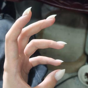 Puede incluir: Una mano con uñas acrílicas blancas, largas y puntiagudas. Las uñas tienen forma de garras, extendiéndose más allá de las puntas de los dedos. El tono de piel es claro y los dedos están ligeramente curvados.