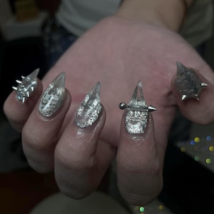 May include: Close-up of a hand with long, clear acrylic nails. Some nails feature silver glitter, while others have silver spikes and a barbell piercing. The nails are pointed and have a glossy finish.