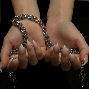 May include: A silver chain necklace held between two hands with long, pointed, white nails. The chain is made of thick, interlocking links. The background is black, creating a contrast with the bright chain.