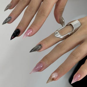May include: Close-up of a hand with long, pointed, gel nails in various designs. The nails feature colors like black, gray, and pink, with a gradient effect. A silver ring with an open rectangular design is also visible.