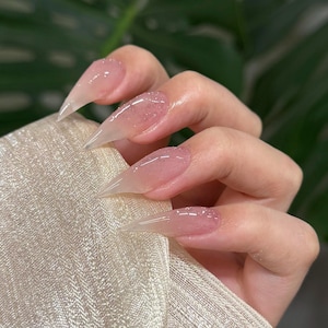 Puede incluir: Primer plano de una mano con uñas acrílicas largas, puntiagudas y transparentes. Las uñas tienen un sutil tono rosado con acentos de purpurina, creando una apariencia delicada y elegante. La mano descansa sobre una tela clara y texturizada.