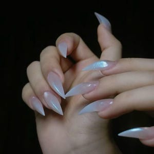 May include: A hand with long, pointed, iridescent acrylic nails. The nails are a pale, translucent color with a subtle shimmer, and are shaped into sharp points. The background is a dark, solid color.