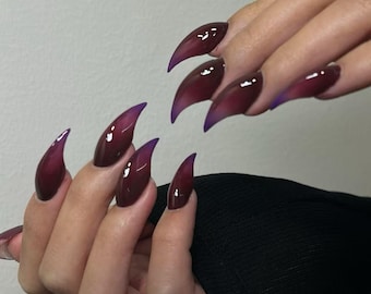 May include: A hand with long, pointed, burgundy-colored acrylic nails with a purple ombre effect at the tips. The nails are glossy and reflect light, creating a dramatic and stylish look. The nails are a deep, rich color.