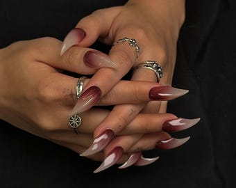 May include: Close-up of long, pointed acrylic nails with a gradient design. The nails transition from a clear base to a deep burgundy color at the tips. Silver rings adorn the fingers, adding a touch of sparkle. The nails are a fashionable accessory.