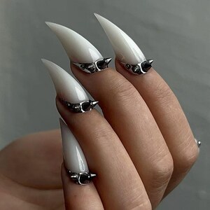 Puede incluir: Primer plano de una mano con uñas largas, blancas y puntiagudas. Cada uña tiene un anillo plateado con una pequeña espiga negra. Las uñas son de color blanco brillante, y los anillos añaden un toque de estilo atrevido.