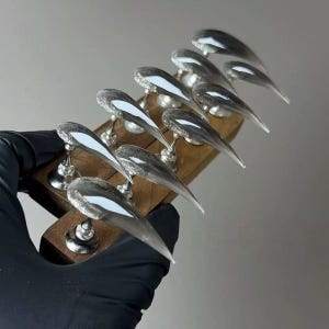 Puede incluir: Un conjunto de uñas acrílicas largas y transparentes con forma puntiaguda, cada una con una base plateada y una franja blanca. Las uñas se exhiben en un soporte de madera con soportes plateados, mostrando un diseño moderno de arte de uñas.