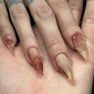May include: Long, pointed stiletto-shaped artificial nails featuring a translucent, glossy finish with blended streaks of deep reddish-brown and muted beige tones, creating a marbled, smoky effect across the surface of each nail extension.
