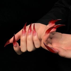 May include: Close-up of hands with long, sharp, red and clear acrylic nails. The nails are shaped into a pointed, claw-like design.
