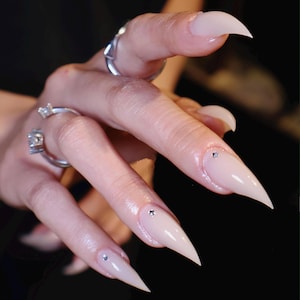 May include: Close-up of long, almond-shaped, light pink nails with a glossy finish. Each nail has a small, sparkling rhinestone near the cuticle. Silver rings adorn the fingers, adding a touch of elegance to the manicure.