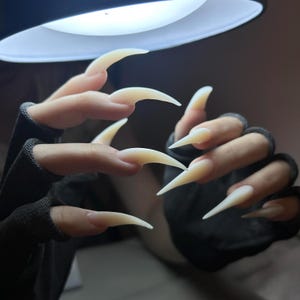 Może przedstawiać: Zbliżenie na długie, spiczaste, jasne sztuczne paznokcie. Paznokcie mają kształt pazurów, wystających z palców. Dłonie noszą ciemnoszare rękawiczki bez palców, z jasnym światłem w tle.