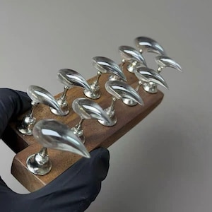 May include: A collection of ten silver-toned, teardrop-shaped decorative objects mounted on a rectangular wooden base. Each object has a slender stem and a rounded base. The objects are arranged in two rows.