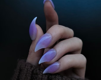 May include: Close-up of a hand with long, pointed, light purple and white ombre nails. The nails are shaped like claws and have a shimmery finish.
